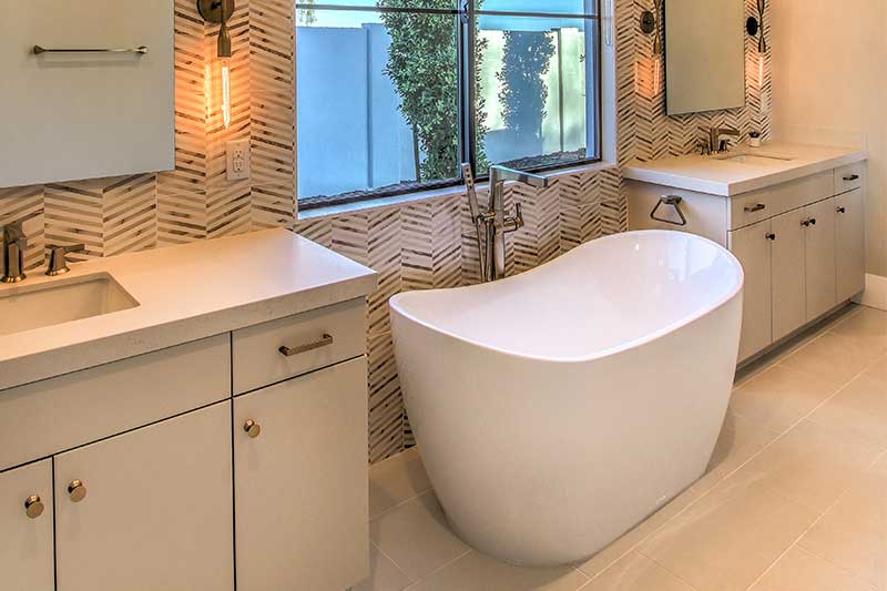A modern bathroom with a freestanding bathtub, dual vanities, geometric wall tiles, and a large window.