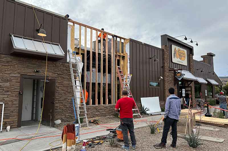 Construction workers repairing the exterior of a commercial building with ladders and tools. Signs indicate businesses inside.