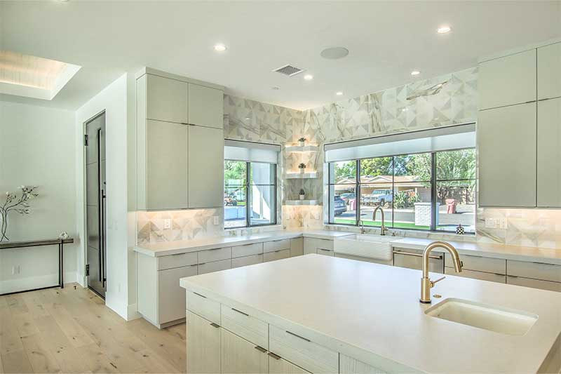 Modern kitchen with light wood floors, white cabinets, and a large island. Features include geometric backsplash, large windows, and gold faucet fixtures.