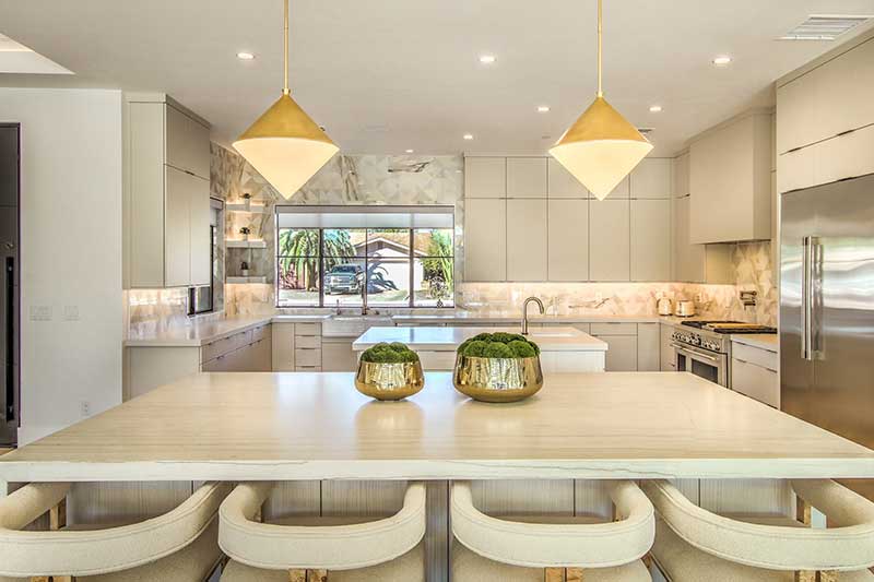 Modern kitchen with beige cabinets, marble countertops, island with beige stools, two pendant lights, and a stainless steel refrigerator.