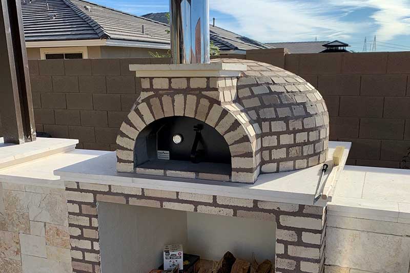 Brick outdoor pizza oven with a dome shape and chimney on a stone counter, with firewood stored below.