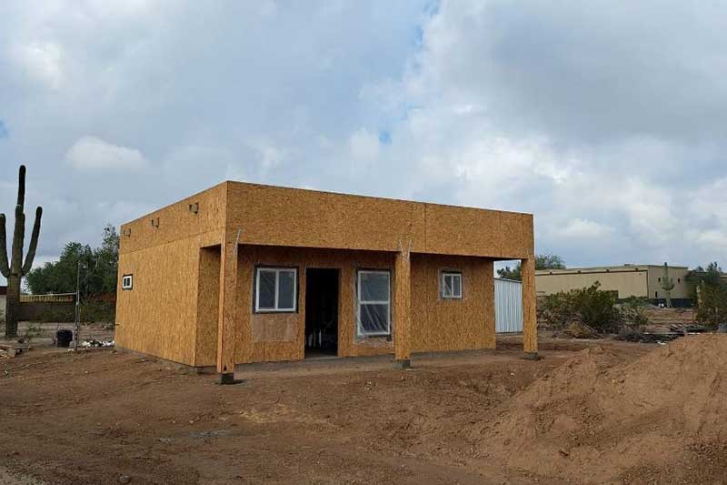 A partially constructed building with a flat roof stands on a dirt lot, surrounded by other structures and a few cacti under a cloudy sky.